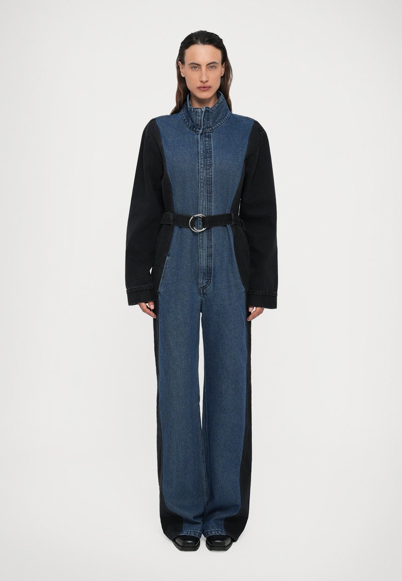 Salopette in denim con corpetto aderente, colletto alto, maniche lunghe nere, gambe larghe e una cintura nera con fibbia circolare. Design bicolore.