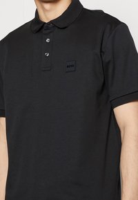 Svart polo skjorta i bomull, med en tvåknappsslutning, ribbad krage och ett litet svart logopatch på bröstet.