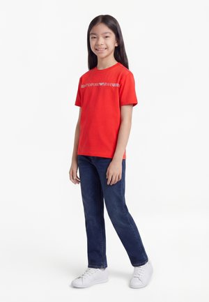 Ragazza giovane in piedi, sorridente, indossa una t-shirt rossa Emporio Armani, jeans blu e sneakers bianche su uno sfondo bianco uniforme.