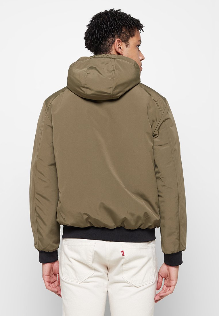 Giacca imbottita verde oliva con cappuccio, polsini e orlo elastici. Texture liscia con un design aerodinamico, caratterizzata da un dettaglio alla cucitura posteriore.