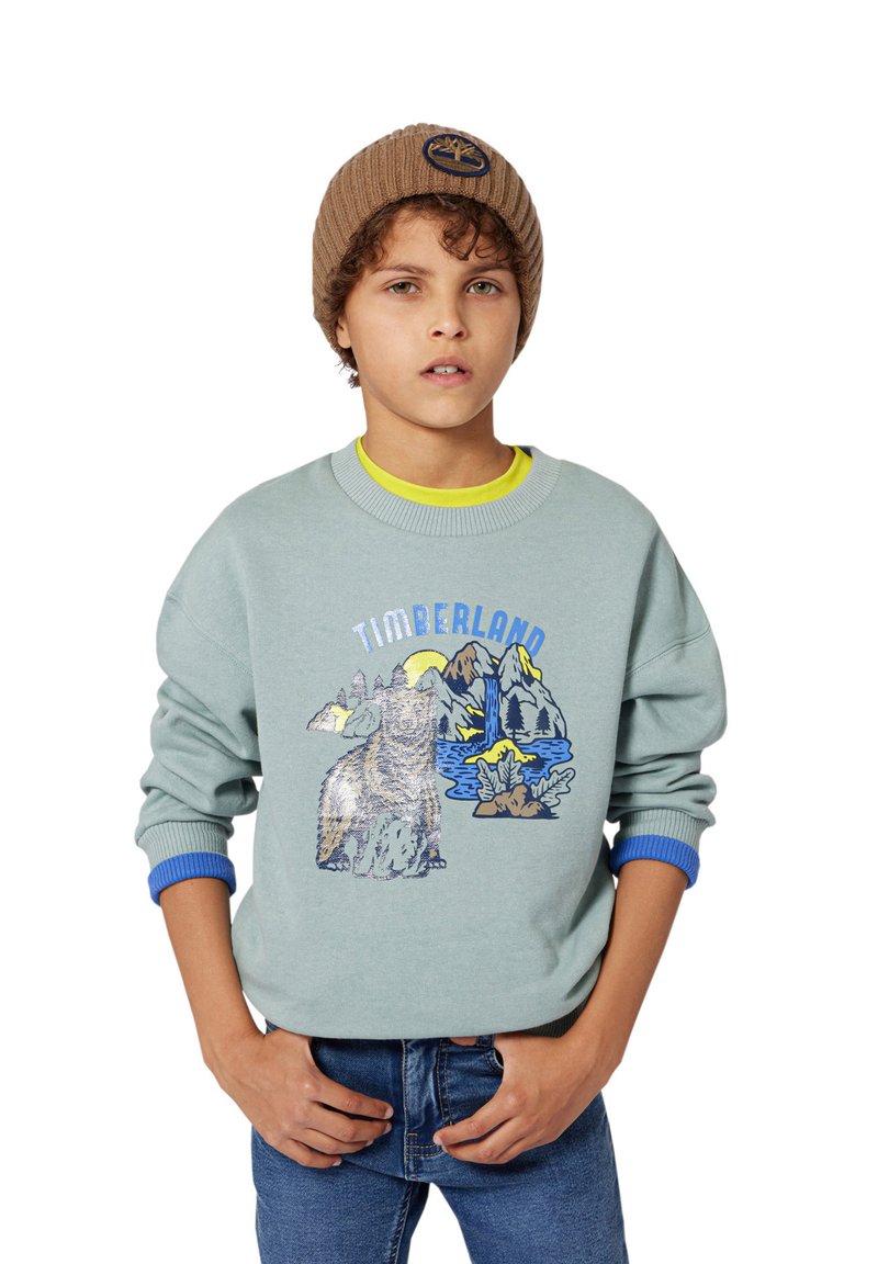 Jasnoniebieski sweatshirt z graficznym motywem niedźwiedzia i gór, z ryflowanymi mankietami w kolorze niebieskim, noszony na żółtej koszulce, w połączeniu z niebieskimi dżinsami.