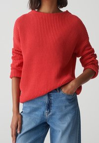 Person trägt einen leuchtend roten, gerippten Strickpullover mit hochgerollten Ärmeln und hellblauen, hoch taillierten Jeans, eine Hand in der Tasche.