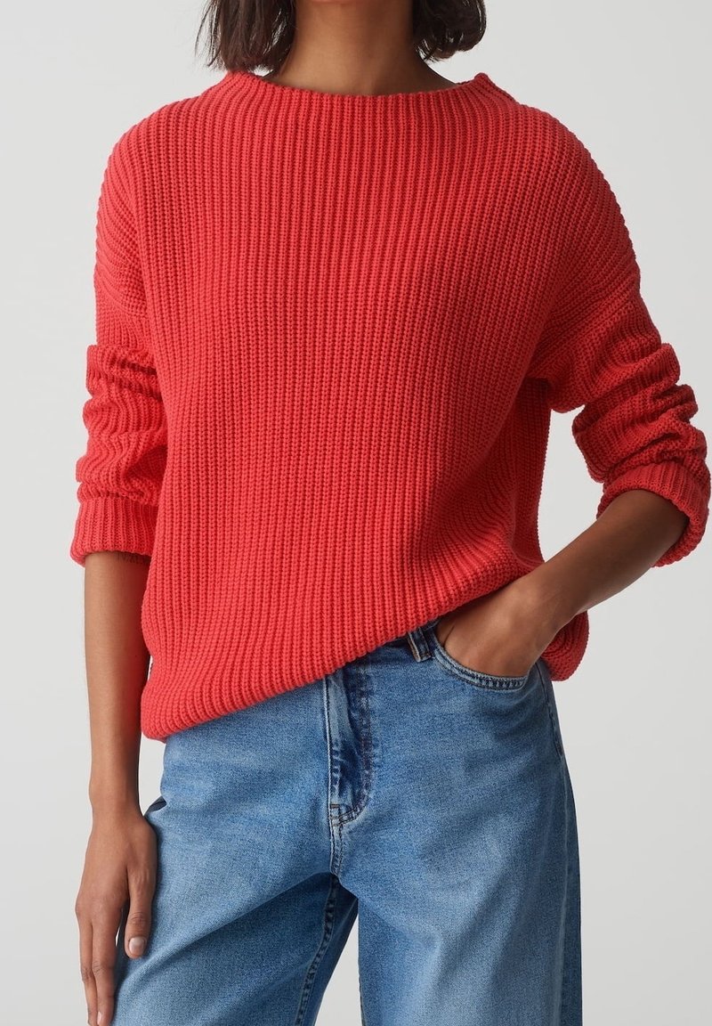 Person trägt einen leuchtend roten, gerippten Strickpullover mit hochgerollten Ärmeln und hellblauen, hoch taillierten Jeans, eine Hand in der Tasche.