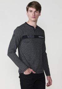 Majica s dugim rukavima, prugasta, crna i siva, s Henley izrezom i crnim platnenim metkom s gumbima i logotipom na prsima.