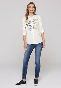 Camicia bianca a maniche lunghe con testo stampato multicolore, abbinata a jeans blu aderenti e sneakers nere. Design con bottoni e maniche arrotolate.