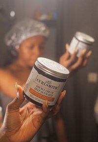 Curlsmith Double Cream Deep Quencher in een zwarte pot met een zilveren deksel, voorzien van oranje en zwarte tekst, vastgehouden door een hand met lange nagels.