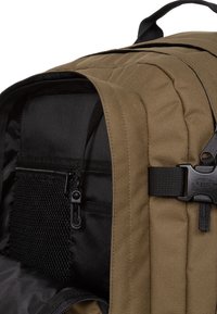 Sac à dos en tissu beige avec un intérieur noir, comprenant une poche zippée, un compartiment en mesh et des attaches de sangle réglables. Détail de couture horizontale.