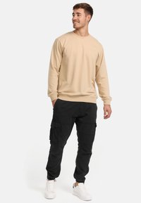 Felpa beige con maniche lunghe e polsini a coste, abbinata a pantaloni cargo neri con molteplici tasche e polsini elastici alle caviglie, indossata con sneakers bianche.