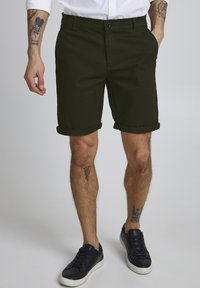 Shorts en coton vert foncé avec des poignets retroussés, un bouton à l'avant et des poches sur les côtés. Associés à des baskets noires.