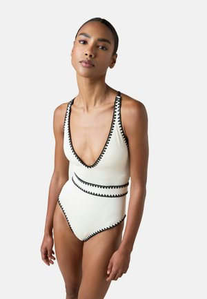 Kobieta w białym teksturowanym jednoczęściowym kostiumie kąpielowym z czarną zygzakowatą lamówką i głębokim dekoltem w kształcie litery V, stojąca na jednolitym jasnym tle.