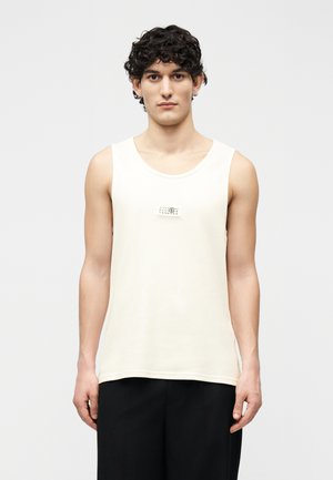 Junger Mann mit dunklem, lockigem Haar trägt ein ärmelloses beiges Tanktop und schwarze Hosen, steht vor einem schlichten hellen Hintergrund.