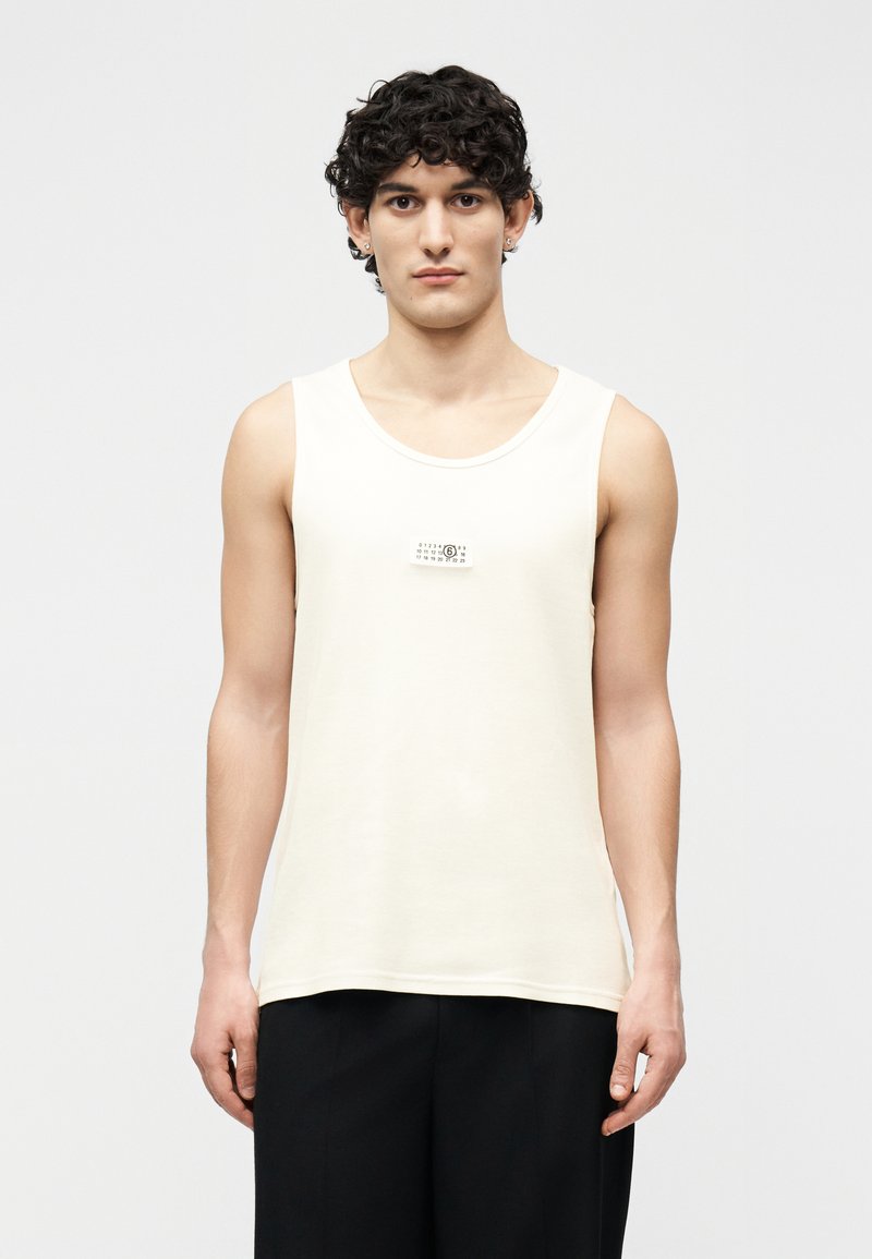 Νεαρός άνδρας με σκουρόχρωμα, σγουρά μαλλιά φοράει αμάνικη κρεμ μπλούζα και μαύρο παντελόνι, στέκεται μπροστά σε απλό ανοιχτό φόντο.