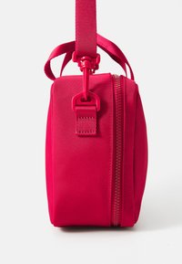 Sac en tissu rouge avec une texture lisse, de forme rectangulaire, doté d'une poignée supérieure et d'une fermeture éclair. Il présente une sangle et des éléments en matériel assortis de couleur rose.