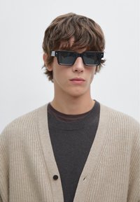 Zwarte rechthoekige zonnebril met een dik montuur, gedragen over een donkergrijs shirt en een beige geribbelde cardigan met een knoopdetail.