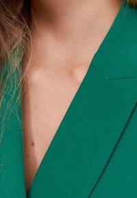 Veste verte avec un décolleté en V profond et des revers aiguisés, confectionnée dans un tissu texturé. Gros plan mettant en valeur les détails de la couture.