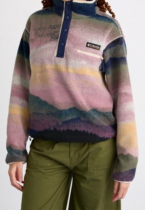 Personne portant un pull polaire multicolore Columbia avec un col boutonné et un pantalon vert olive, montré des épaules jusqu'à mi-cuisse.