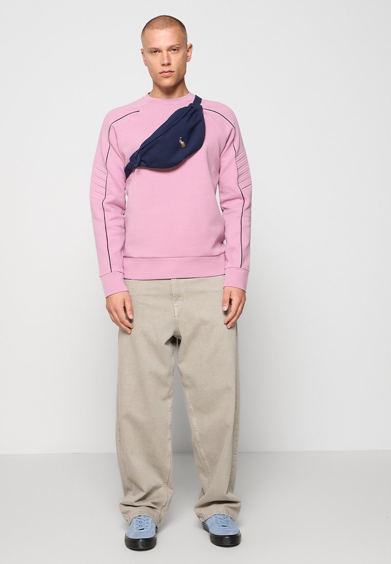 Sweatshirt rose avec des détails côtelés sur les manches, associé à un pantalon beige ample et des baskets bleu clair. Un sac à taille bleu foncé est porté sur la poitrine.