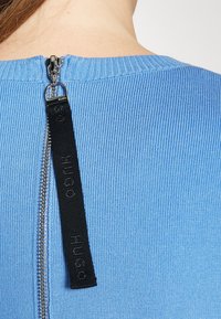 Haut en tissu tricoté côtelé bleu, avec une fermeture éclair argentée au dos et une languette en tissu noir embossée du mot "Hugo".