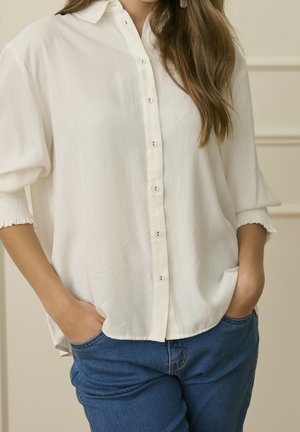 Kobieta ubrana w luźną białą koszulę na guziki z podwiniętymi rękawami i niebieskie dżinsy, ze swobodnie włożonymi rękami do kieszeni, stojąca w pomieszczeniu.