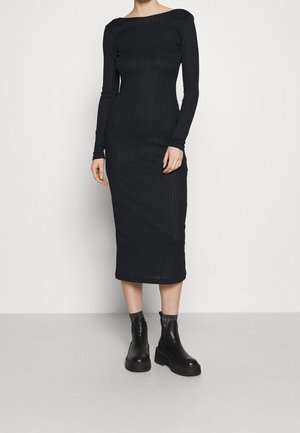 Kobieta ubrana w czarną, prążkowaną sukienkę midi z długim rękawem oraz czarne botki za kostkę, stojąca na tle jednolitej białej ściany.