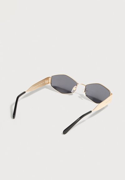 Εξαγωνικά γυαλιά ηλίου με σκούρους φακούς και χρυσό μεταλλικό σκελετό, με μαύρες καμπύλες άκρες στα πλαϊνά, πάνω σε λευκή επιφάνεια.