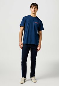 Tummansininen pyöreäkauluksinen T-paita, jossa on punainen "Wrangler"-logo, yhdistettynä tummiin farkkuihin ja beigeihin lenkkareihin. Yksinkertainen muotoilu, rento istuvuus.