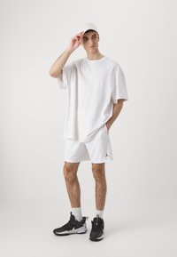 Fehér, oversize póló, fehér hálós rövidnadrág fekete logóval és fekete sneaker. Világos sapka van rajta, a kezét a homlokára tette.