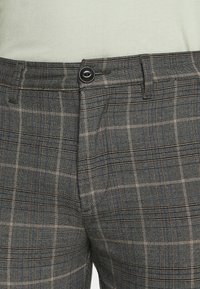 Grå rutiga byxor i lättviktstyg, med ett blått och brunt rutmönster. Inkluderar en svart knapp och sidofickor.