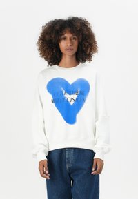 Felpa bianca con una grande grafica a forma di cuore blu e testo nero che recita "L'AMORE È UN SENTIMENTO NON UNA DECISIONE." Vestibilità ampia e texture morbida.