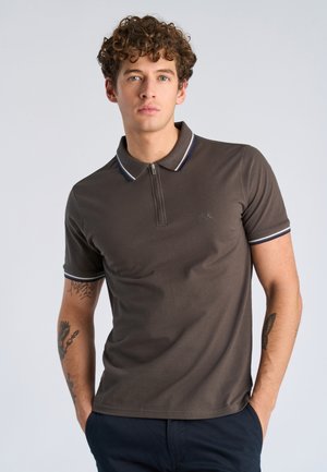 Jonge man met krullend haar draagt een bruin poloshirt met korte mouwen, gestreepte kraag en manchetten, staand met handen in de zakken van donkere broek.