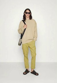 Sweatshirt bege, calças cargo em tom de oliva e sandálias pretas. Acessório: bolsa de ombro preta e óculos de sol. Conjunto casual com um corte descontraído.