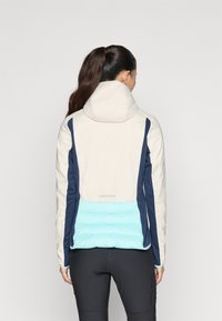 Opgezette gewatteerde jas met een beige hoofdkleur, marineblauwe zijpanelen en een lichtblauwe onderkant. Glad oppervlak met een modern ontwerp en logo-detail.