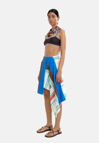 Brązowy top na krótki rękaw z niebieskimi wykończeniami, połączony z wielokolorową spódnicą w paski, w której dominują niebieskie, zielone i czerwone akcenty. Całość uzupełniają sandały.