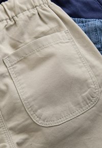 Beige stofshorts med elastisk talje, der har en syet lappetaske. Teksturen er glat med synlige sømdetaljer.