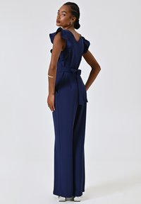 Marineblauer Jumpsuit mit Rüschensäumen, Bindegürtel und geradem Schnitt. Glattes Material mit einem Reißverschluss auf der Rückseite und weit geschnittenen Hosenbeinen.