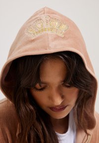 Felpa in velluto rosa cipria con design a corona ricamato in oro e argento. Presenta un cappuccio spazioso e sfoderato, con una lieve lucentezza.