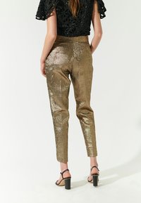 Skinnende gyldne tapered bukser med en tekstureret overflade, parret med en sort blomstret blonde top og sorte blokhælede sandaler.