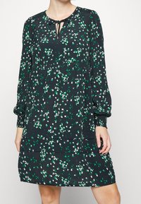 Vestido floral en verde oscuro con patrones en blanco y verde, presenta un escote con lazo y mangas largas con puños elásticos.