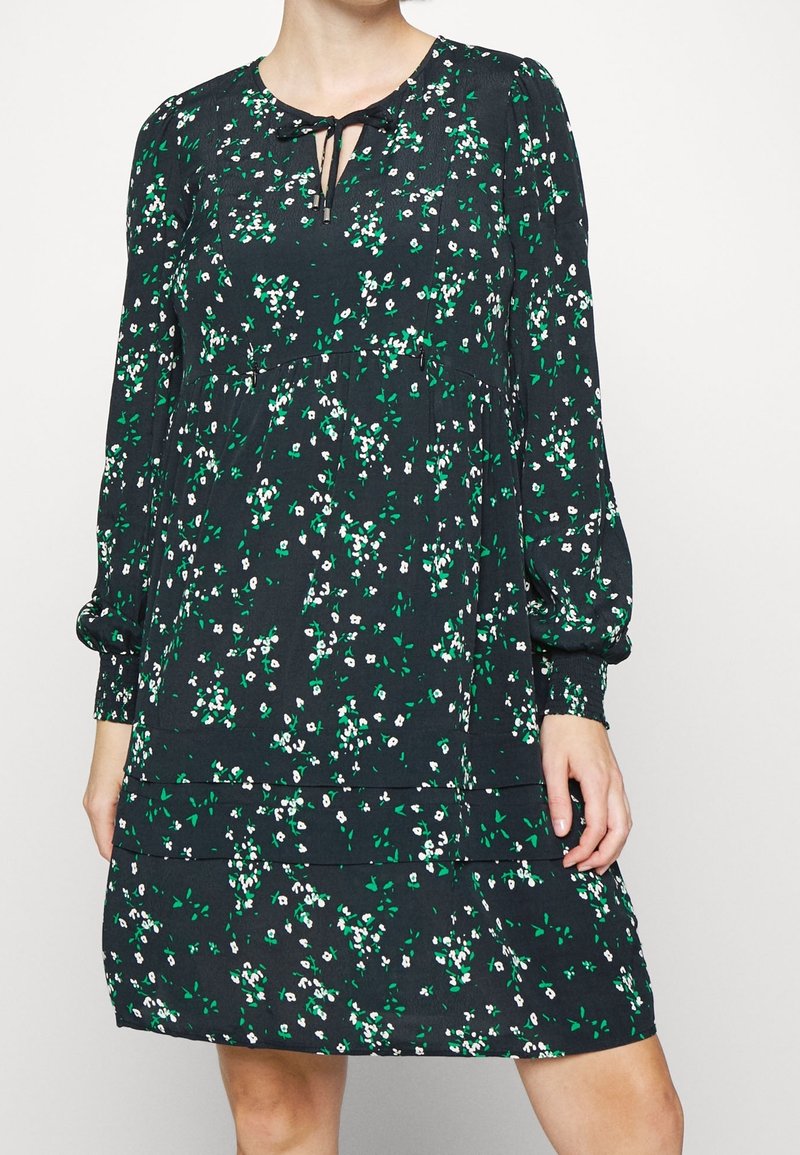 Vestido floral en verde oscuro con patrones en blanco y verde, presenta un escote con lazo y mangas largas con puños elásticos.