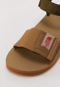 Ruskea sandaali, jossa on teksturoitu pohja, kangasremmit ja pinkki logopatch. Ominaisuuksiin kuuluu litteä muotoilu ja oranssi yksityiskohta pohjan reunalla.