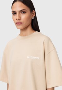 Beżowa koszulka z krótkim rękawem z małym białym logo "ALLSAINTS". Okrągły dekolt i luźny krój. Miękki bawełniany materiał o minimalnej teksturze.