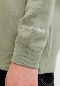Gros plan sur la manche d'un pull en tricot vert clair avec un poignet côtelé et la broderie "Original Studio" près du poignet au-dessus de la main d'une personne.