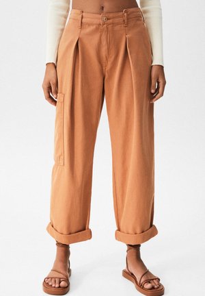 Rotskleurige, wijde broek met hoge taille, zijzak en omgeslagen zoom, gedragen met bruine sandalen met bandjes.