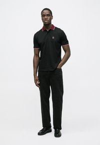 Svart poloshirt med burgunderfarget krage og brodert logo, kombinert med svarte bukser og svarte sko. Modellen står foran en hvit bakgrunn.