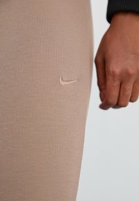 Šviesiai rudos plačios legins su subtiliu Nike logotipu įspaustu ant šlaunies; arti pavaizduota medžiagos tekstūra ir prigludimas.