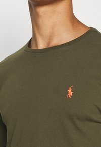 Zbliżenie na osobę noszącą ciemnooliwkową koszulkę z pomarańczowym haftowanym logo gracza polo po lewej stronie klatki piersiowej.