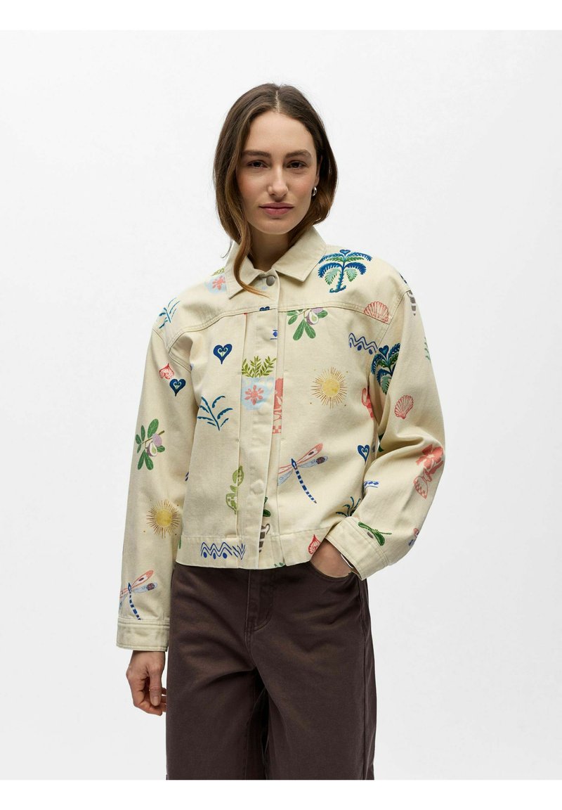 Femme portant une veste beige avec des motifs colorés sur le thème de la nature et un pantalon marron, debout avec une main dans la poche sur un fond uni.