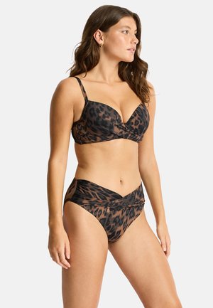 Femme aux cheveux bruns ondulés portant un bikini imprimé léopard marron et noir avec un haut rembourré et un bas taille haute, debout sur un fond clair.