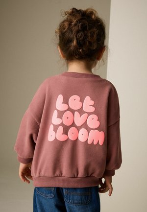Lilla sweatshirt med oversized pasform, der har lyserød boblende tekst "Let Love Bloom" på ryggen. Tekstureret stof med ribstrikkede manchetter.