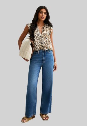Floral ruffled blouse in beige and brown, paired with high-waisted wide-leg blue jeans. Holding a textured, light-colored handbag.